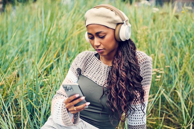 Woman sitting in tall grass wearing cream headphones and using her phone