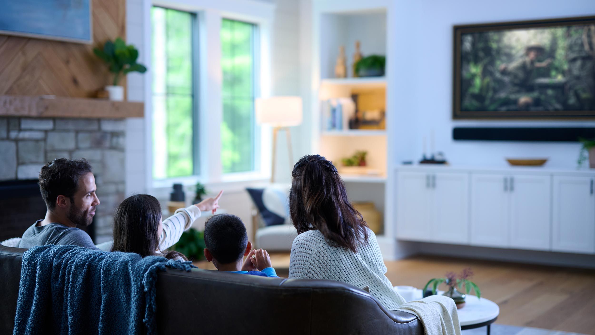 A family gathered in a cozy living room, watching a wall-mounted TV with a soundbar, surrounded by natural light and stylish decor.