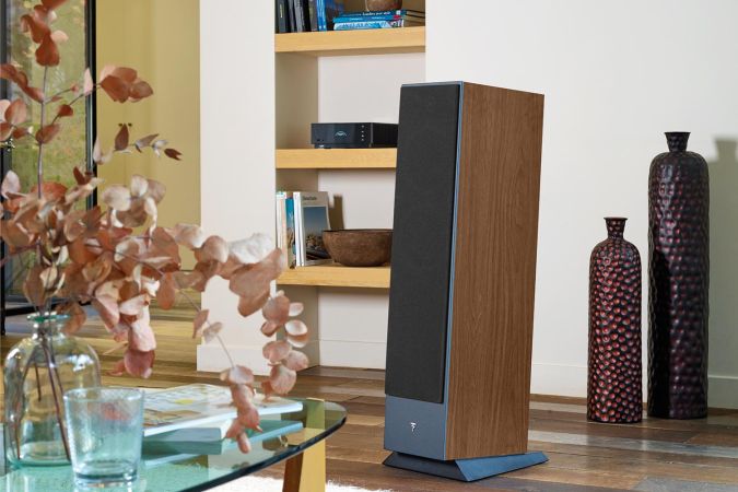 A wooden floor-standing speaker placed in a living room corner, surrounded by neutral decor and soft natural lighting.