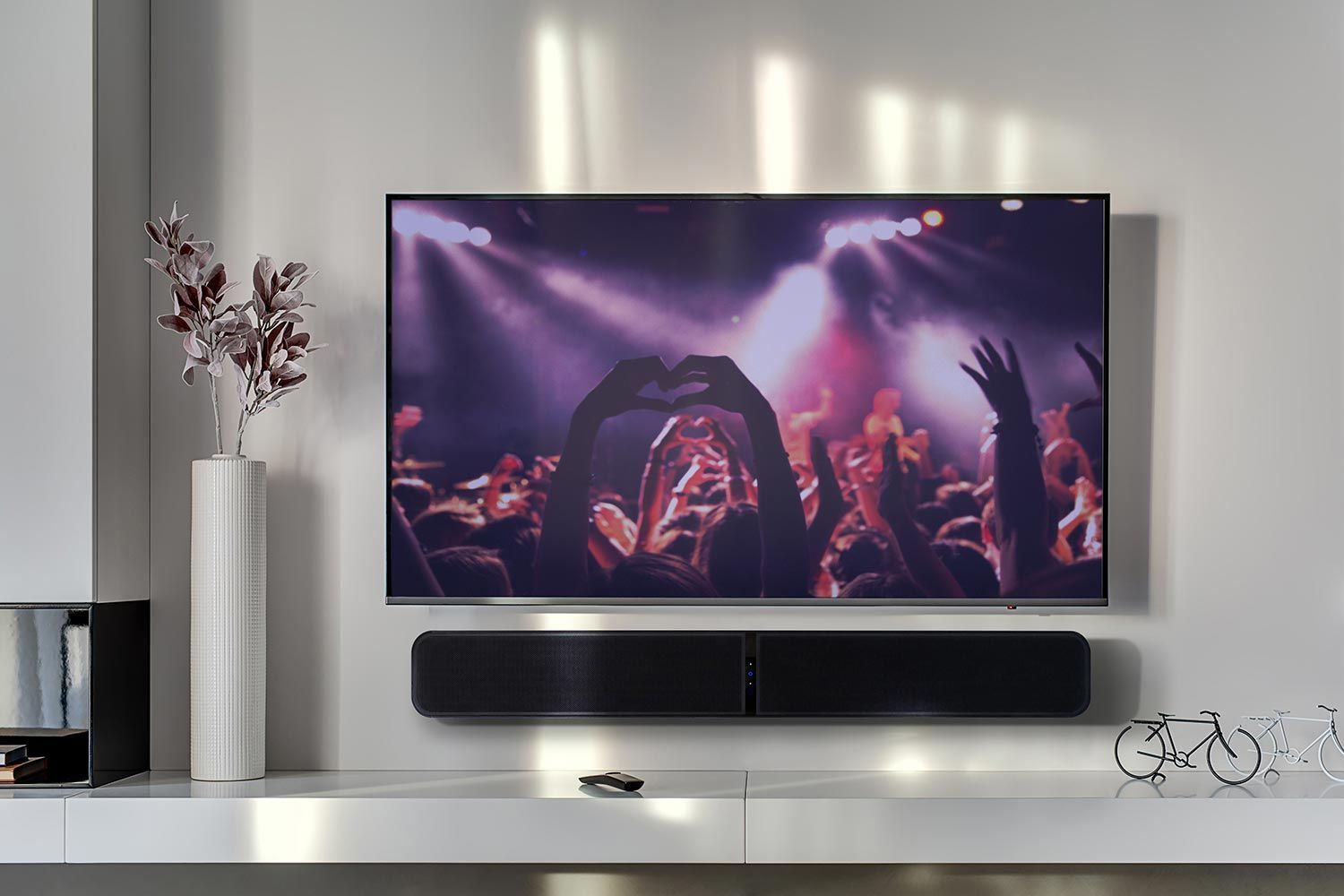 A living room with a mounted TV displaying a concert scene. Below the TV is a Bluesound soundbar, and on a shelf to the left is a tall, white vase with dried leaves. The overall color palette is neutral and contemporary.