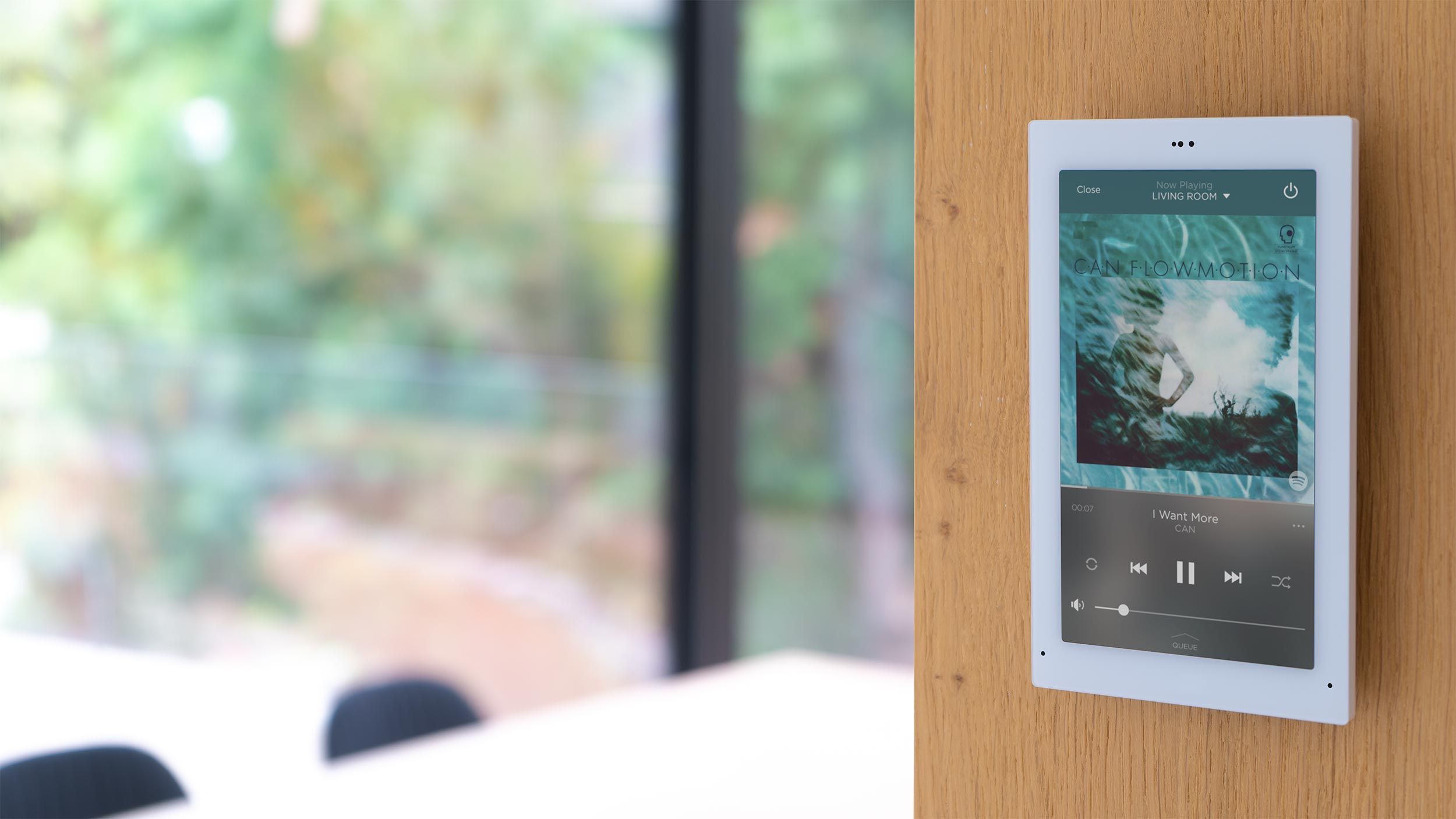 A sleek in-wall touchscreen control panel displaying a music streaming interface, mounted on a wooden surface with a bright, open space in the background.