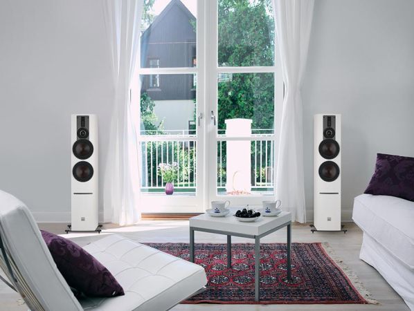 A bright and minimalist living space with white floor-standing speakers, a glass coffee table, and an open balcony view, blending audio quality with elegant design.