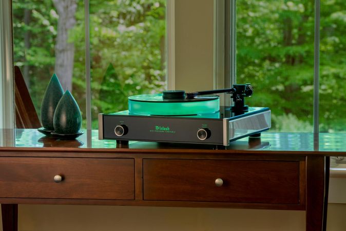 A sleek record player with a glowing green platter on a wooden table in front of a large window overlooking a lush green landscape.